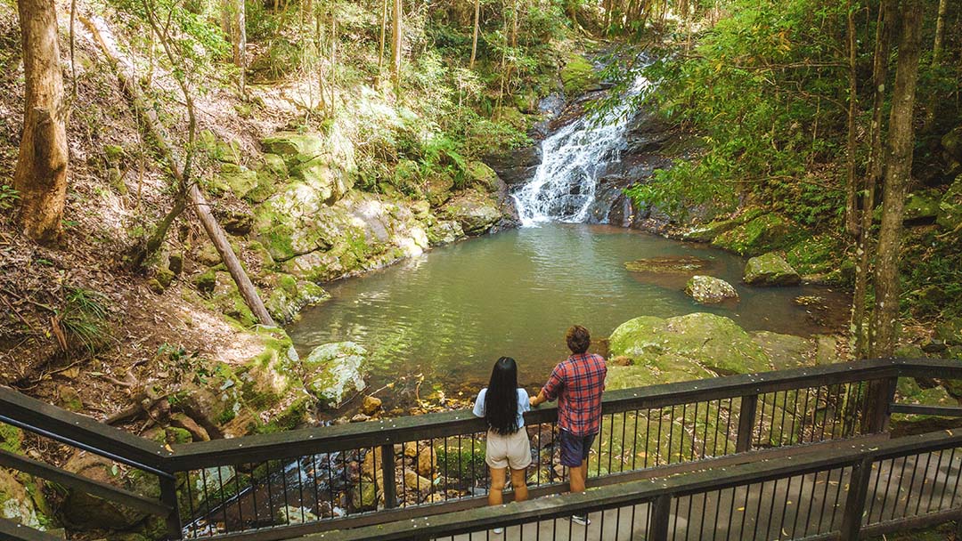 Two People Stand at a Wooden Railing, Looking over a Pool Beneath a Small Forest Waterfall.