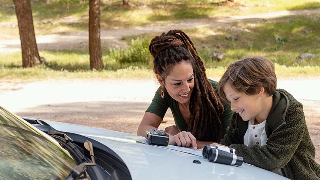 Woman and Boy Leaning over a Car Hood, Examining a Camera and a Pair of Binoculars in an Outdoor Park Setting.
