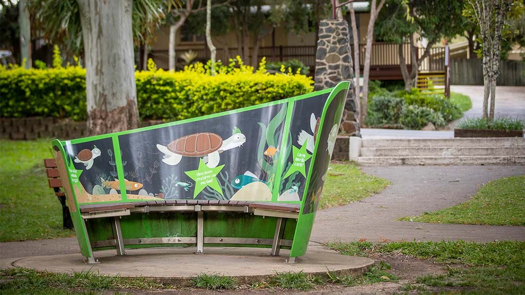 Curved Green Park Bench with Turtle-themed Panels, Set on a Circular Concrete Pad in a Grassy Area.