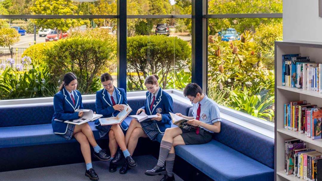 Home_ccps-8-med Copy 1 | Kids in Brisbane, Gold Coast, Sunshine Coast | Kotc Four Students in Blue Blazers and Ties Sit on a Curved Blue Sofa in a Bright Library, Reading Books Together.