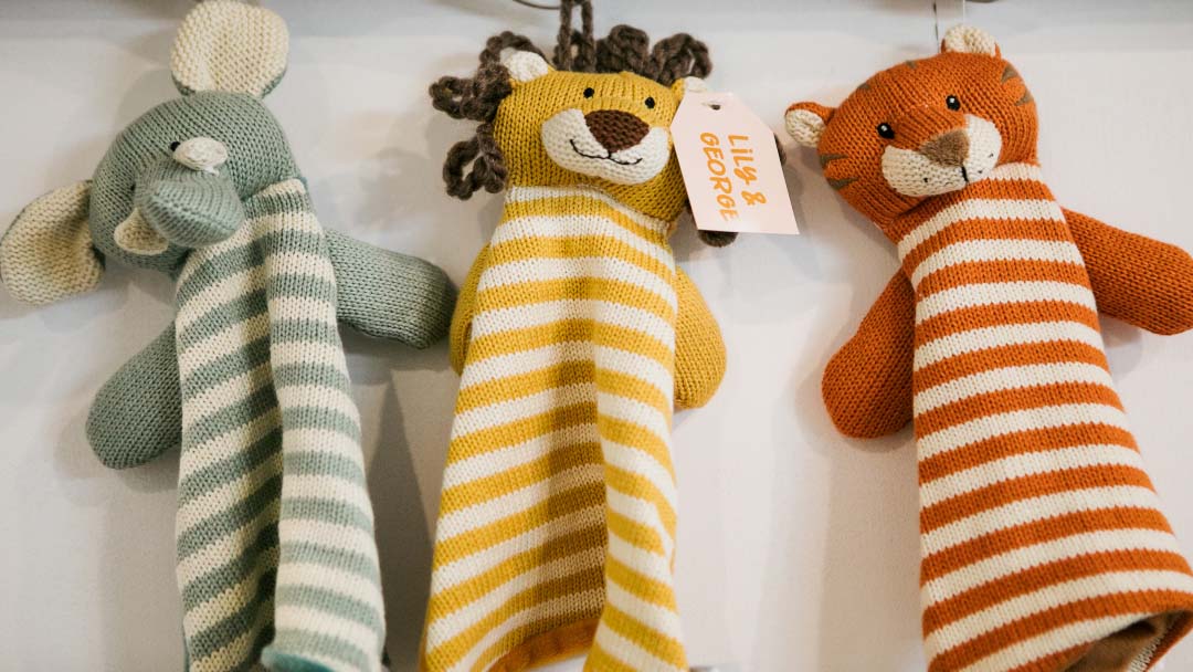 Three Knitted Stuffed Animals with Long Striped Necks in Blue, Yellow, and Orange Hanging on a White Surface; Middle One Has a Tag.