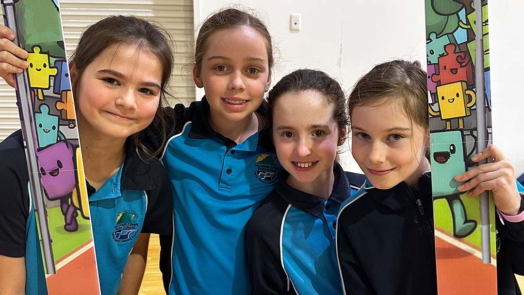 Four Girls in Blue and Black Shirts Pose Together in a Gym, Smiling, with Colorful Puzzle-piece Banners on Both Sides.