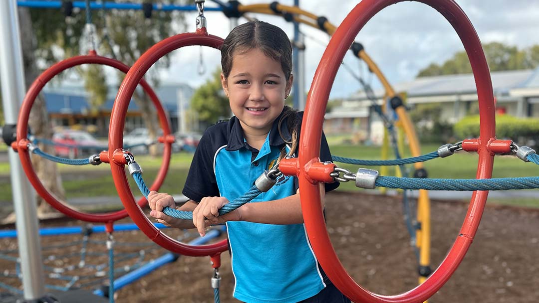 Young Girl in a Blue Shirt Smiles While Standing on Red Circular Climbing Rings at a Playground.