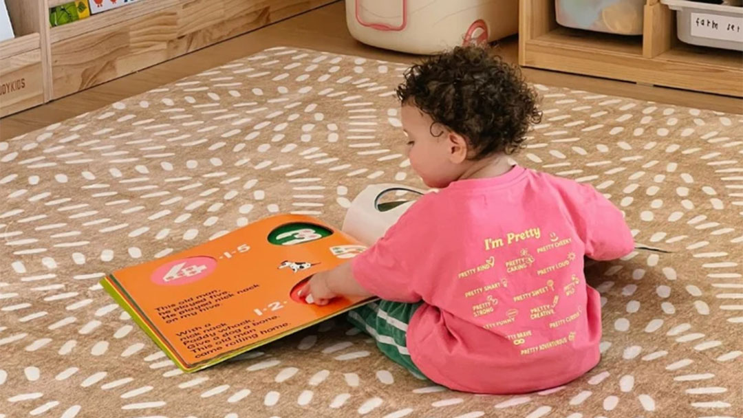 Miss-amara-kids-playmat | Kids in Brisbane, Gold Coast, Sunshine Coast | Kotc Toddler in a Pink Shirt Sits on a Patterned Rug, Reading a Bright Orange Board Book on the Floor.