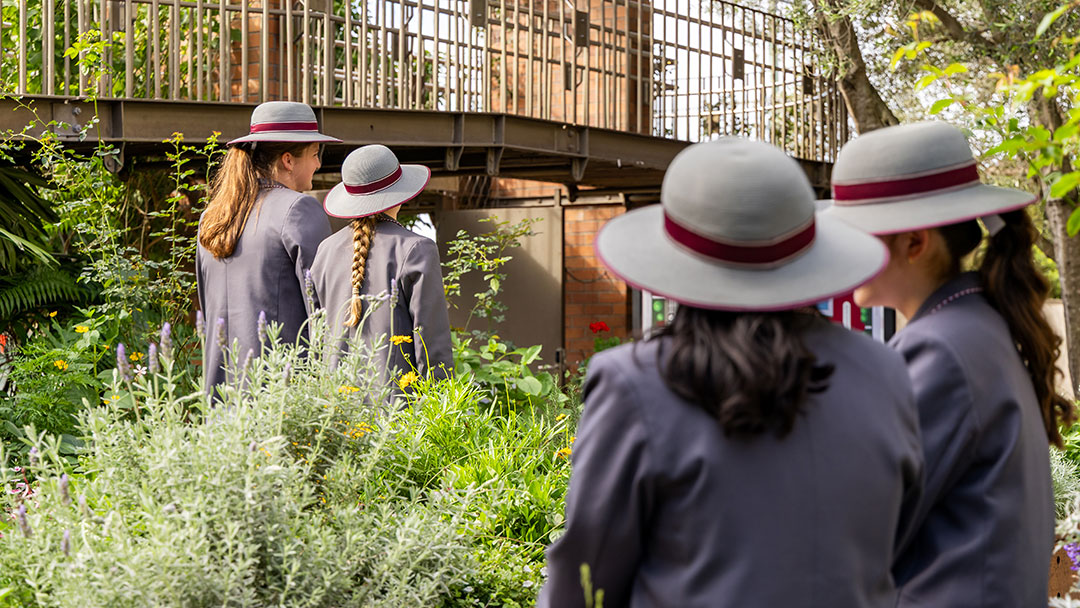 Mount-alvernia-college-gardens---2026 | Kids in Brisbane, Gold Coast, Sunshine Coast | Kotc Four People in Gray Uniforms and White Hats with Red Bands Stand in a Lush Garden Near a Brick Building.