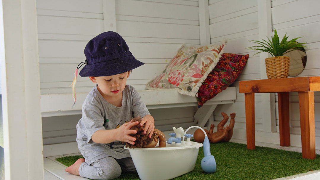Child Wearing a Dark Hat Bathes a Doll in a White Tub on a Sunny Patio Outdoor Play Scene with Bath Toys Nearby