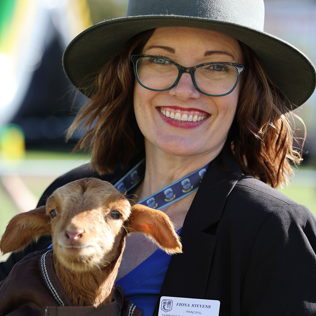 Smiling Woman with Glasses and a Wide-brimmed Hat Holds a Brown Baby Goat Outdoors, Both Looking at the Camera. She Wears a Blazer and Name Badge Visible on Her Chest.
