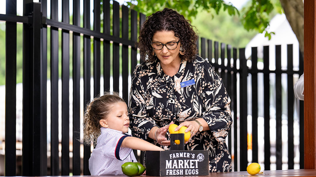The-lakes-college-teacher-with-student | Kids in Brisbane, Gold Coast, Sunshine Coast | Kotc Woman with Glasses and Floral Blouse Helps a Young Girl Pick Lemons at a Farmers Market Stand, Near a Crate Labeled'Farmer's Market Fresh Eggs'