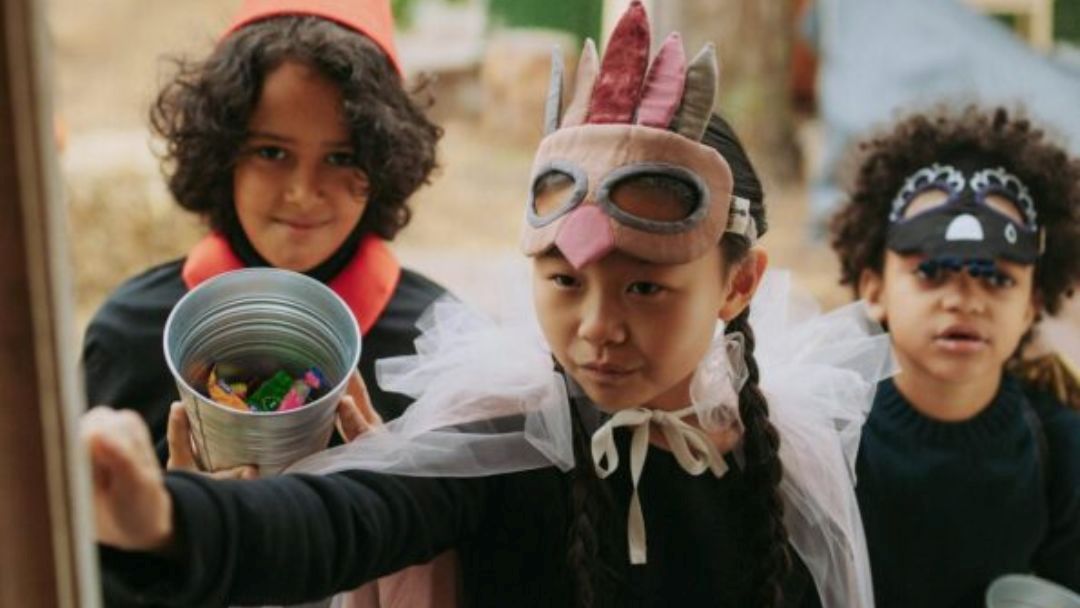 1013_currimundihotel_halloween_hero | Kids in Brisbane, Gold Coast, Sunshine Coast | Kotc Three Kids in Costume Outdoors; Center Girl in a Bunny Mask with Braided Hair, Reaching Forward As Others Watch, Holding a Bucket of Candy.
