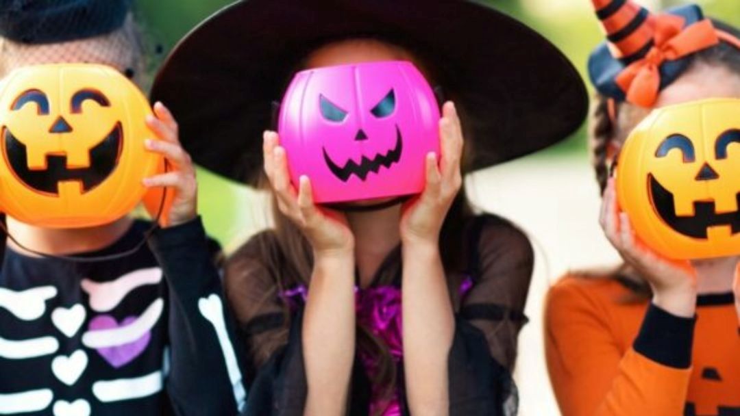 1013_hotelhq_halloween_hero | Kids in Brisbane, Gold Coast, Sunshine Coast | Kotc Three Children in Halloween Costumes Hold Jack-o'-lantern Masks in Front of Their Faces Outdoors, Including a Pink Pumpkin, an Orange Pumpkin, and a Skeleton Outfit Visible on the Left Person.