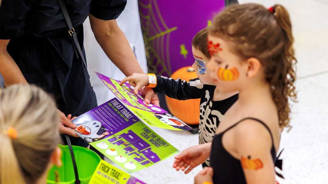 Children in costumes examine a colorful Halloween flyer handed by an adult.