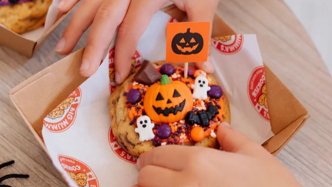 Halloween-themed Cookie Topped with a Jack-o'-lantern Pumpkin, Two White Ghost Figures, a Black Bat, Purple Candies, and Orange Sprinkles in a Takeout Box with a Small Pumpkin Flag