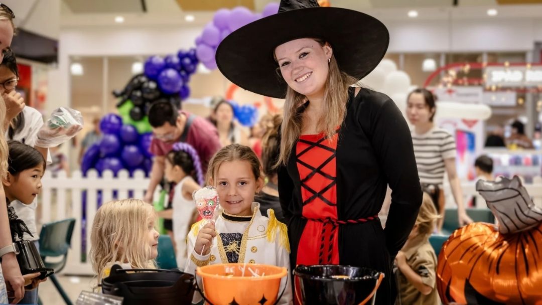 a Young Girl in a White Costume Holds a Pink Lollipop and Smiles Beside a Woman Dressed As a Witch in a Black and Red Outfit at a Bustling Indoor Halloween Event the Mall Center is Lively with Balloons.